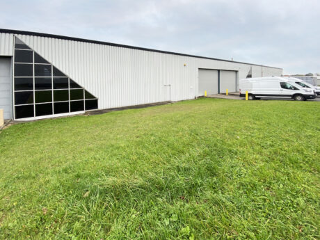 A white industrial warehouse with large windows and garage doors, bordered by a grassy lawn. Several white vans are parked on the right side.