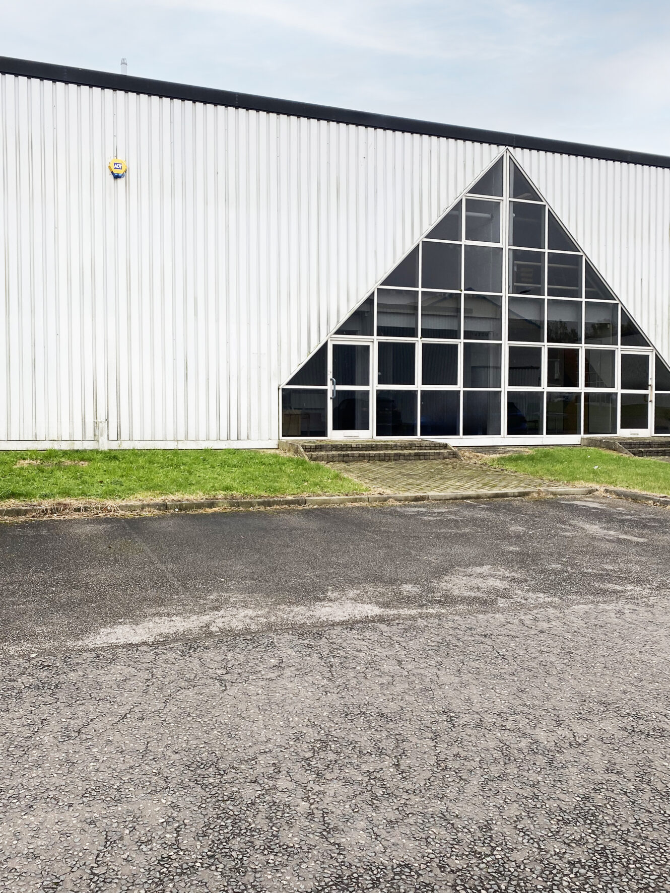A modern industrial building with a triangular glass entrance, corrugated metal siding, and a paved parking area in front.