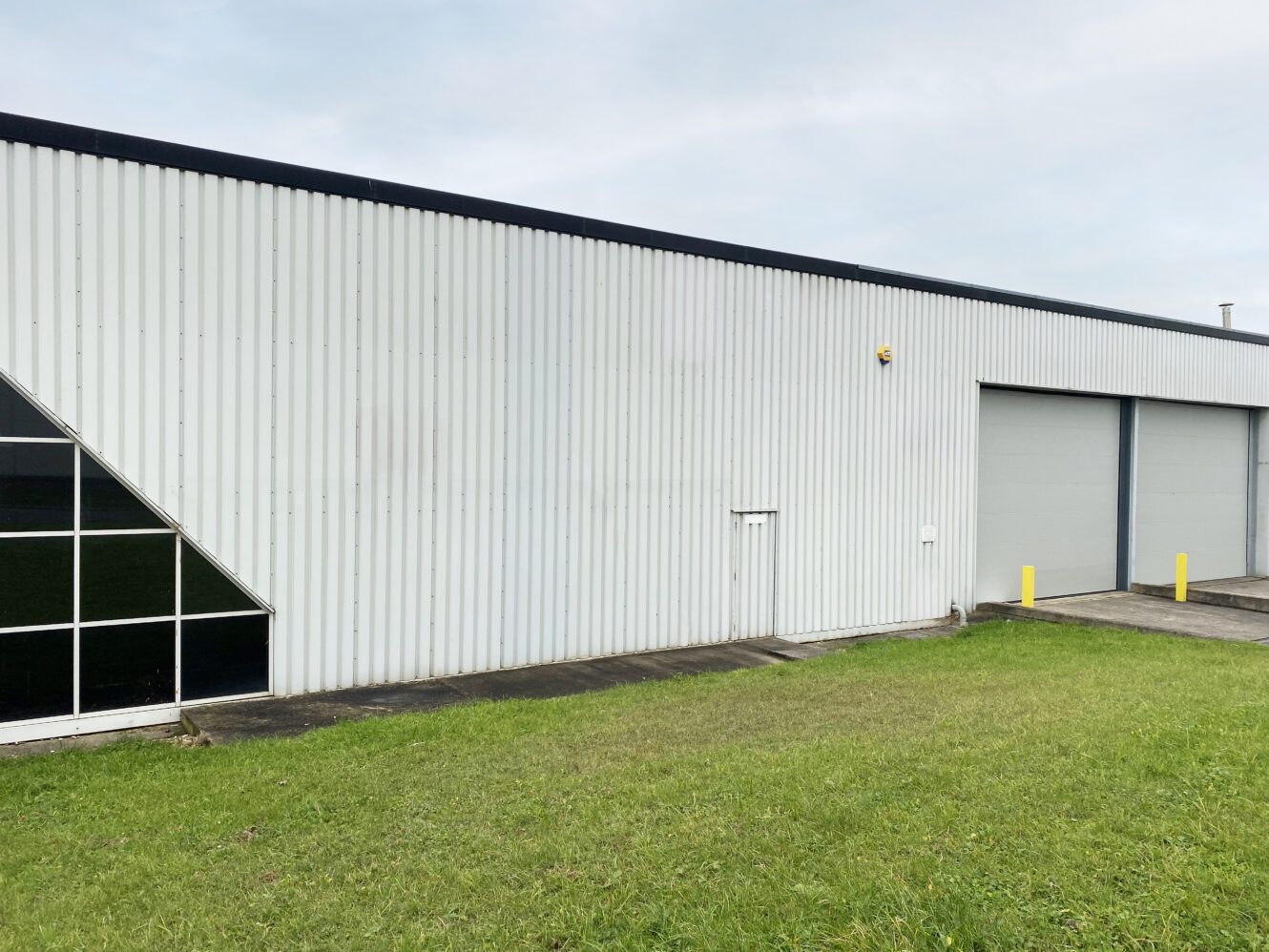 A large industrial building with a metal exterior, a small door, two large garage doors, and a grassy area in the foreground.