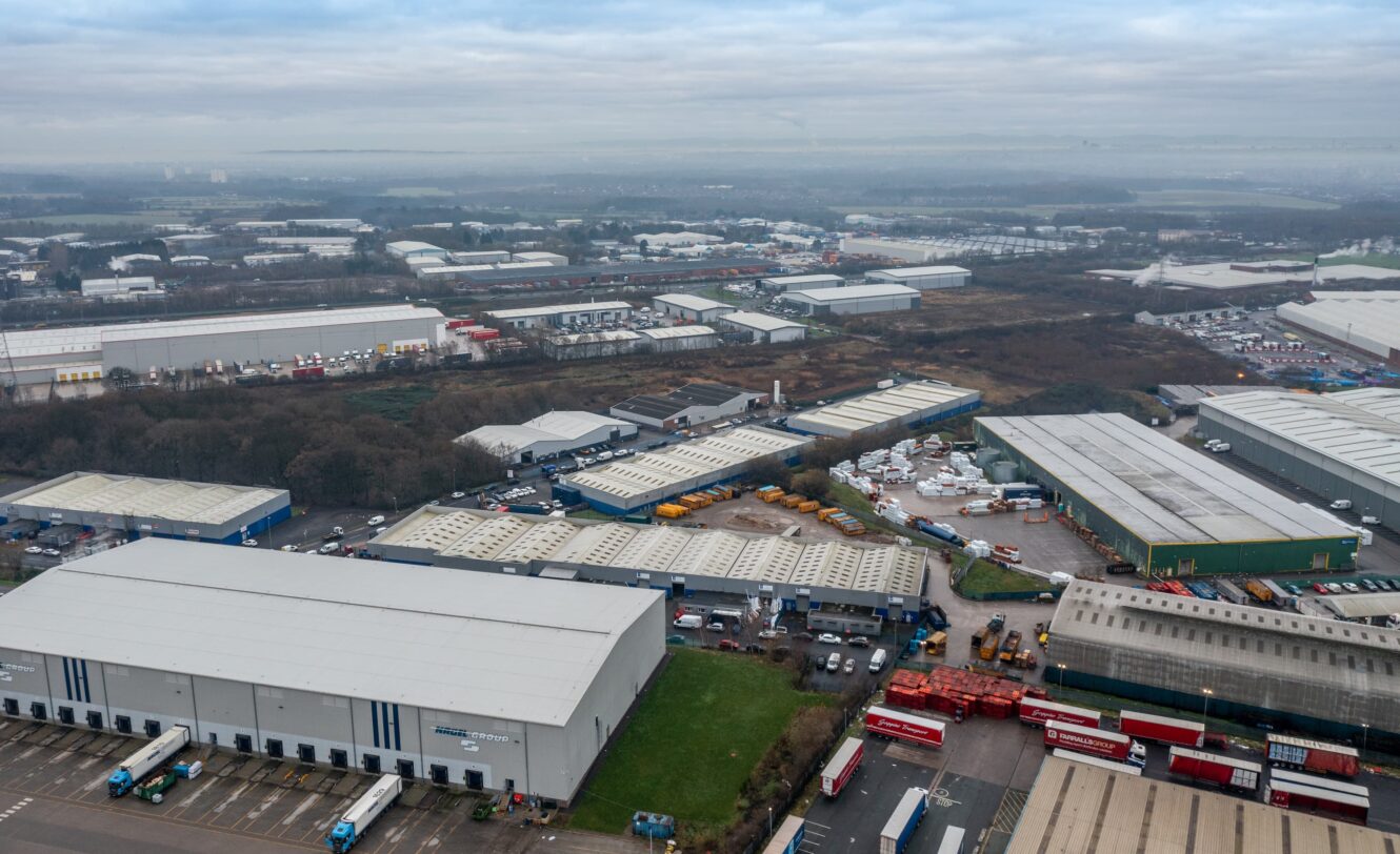 Aerial view of an industrial estate with warehouses, storage yards, parked trucks, and surrounding roads under a cloudy sky.