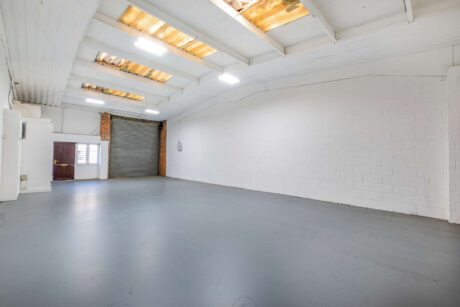Large, empty warehouse with white walls, grey floor, overhead fluorescent lighting, skylights, and a roller shutter door at the far end.