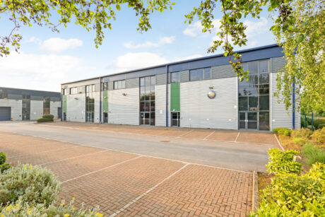 Modern industrial or office building with large windows and metal siding, surrounded by greenery and an empty brick-paved parking lot.