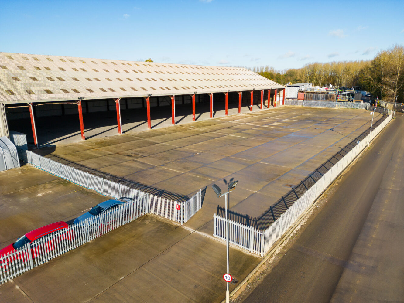 A large, empty, fenced industrial lot with a covered storage area, adjacent to a road and bordered by trees under a clear sky.