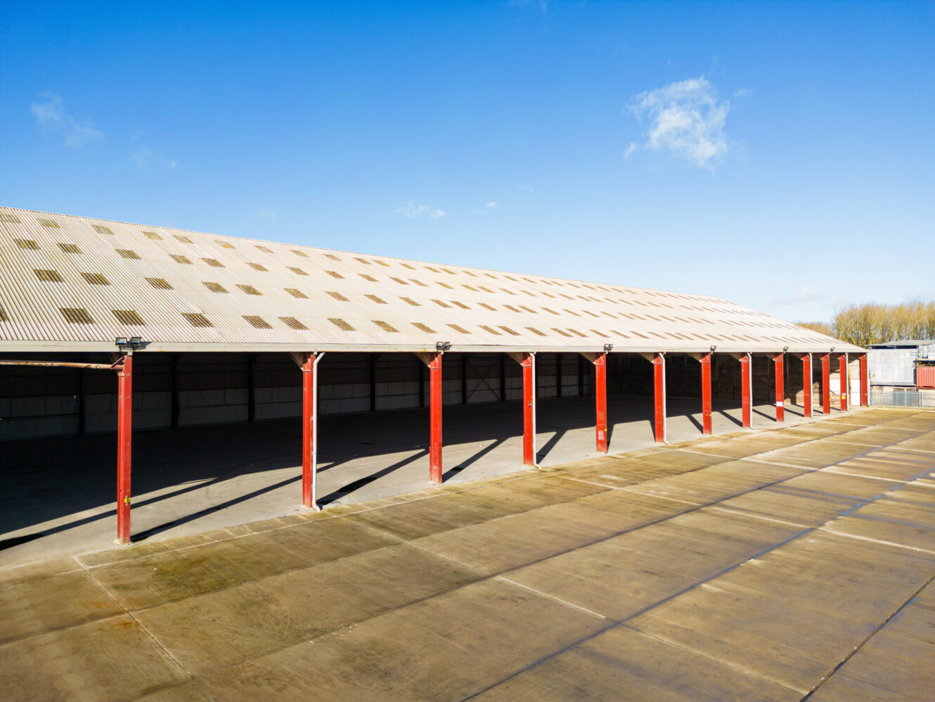 Large open-sided warehouse with a corrugated metal roof, supported by red steel columns, on a concrete surface under a clear blue sky.