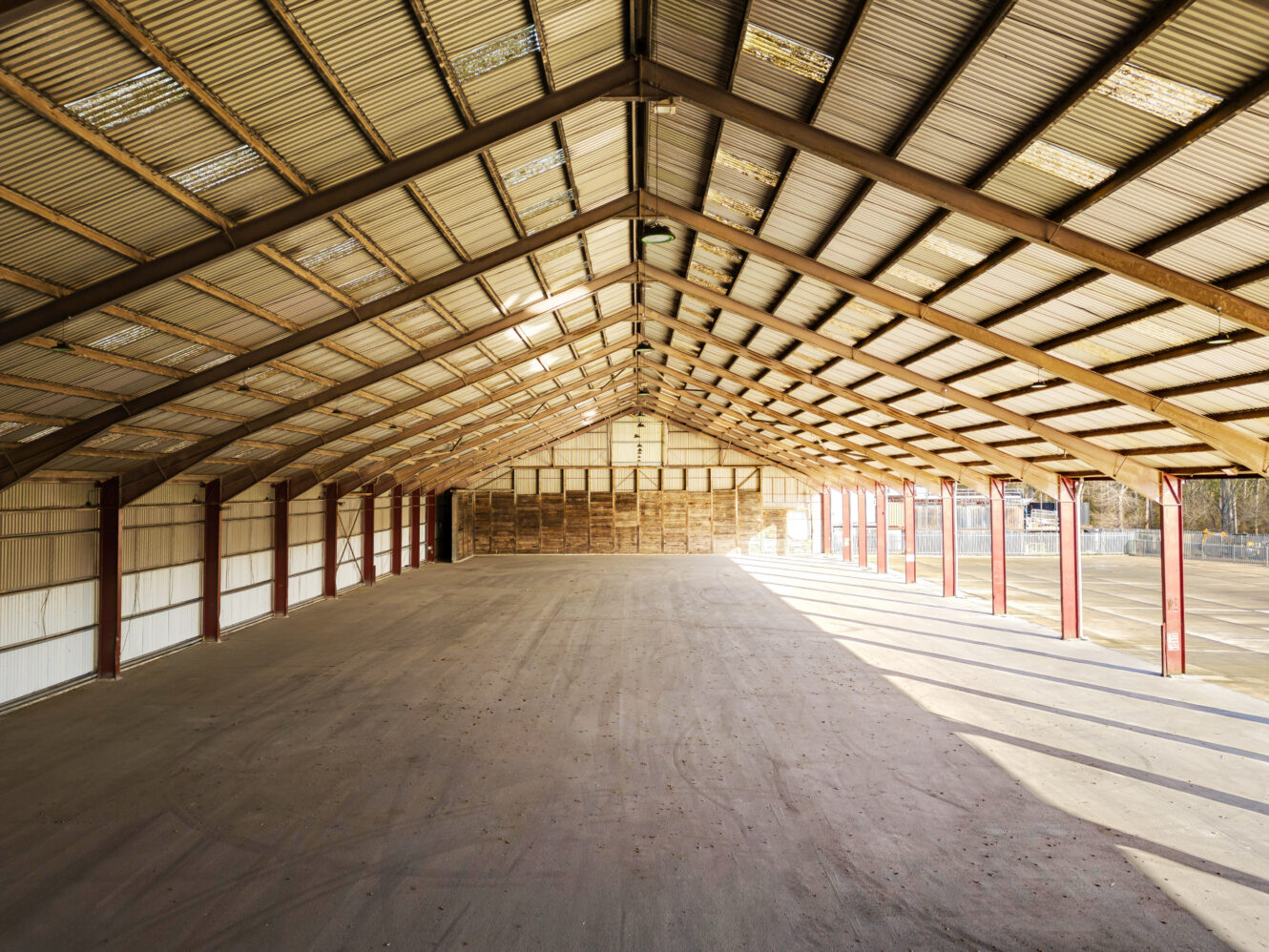 Interior view of an empty, large warehouse with a metal roof, exposed beams, and sunlight streaming in from the right side.