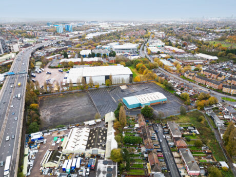 Aerial view of an industrial area with warehouses, parking lots, residential houses, and a major highway on the left side of the image. Trees with autumn foliage are scattered throughout.