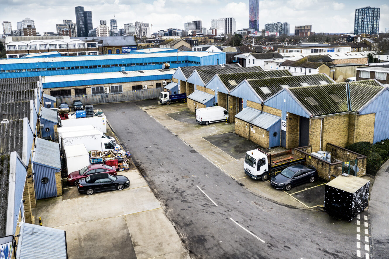 Aerial view of an industrial estate with blue-roofed warehouses, parked vans and cars, and high-rise buildings visible in the distant background.