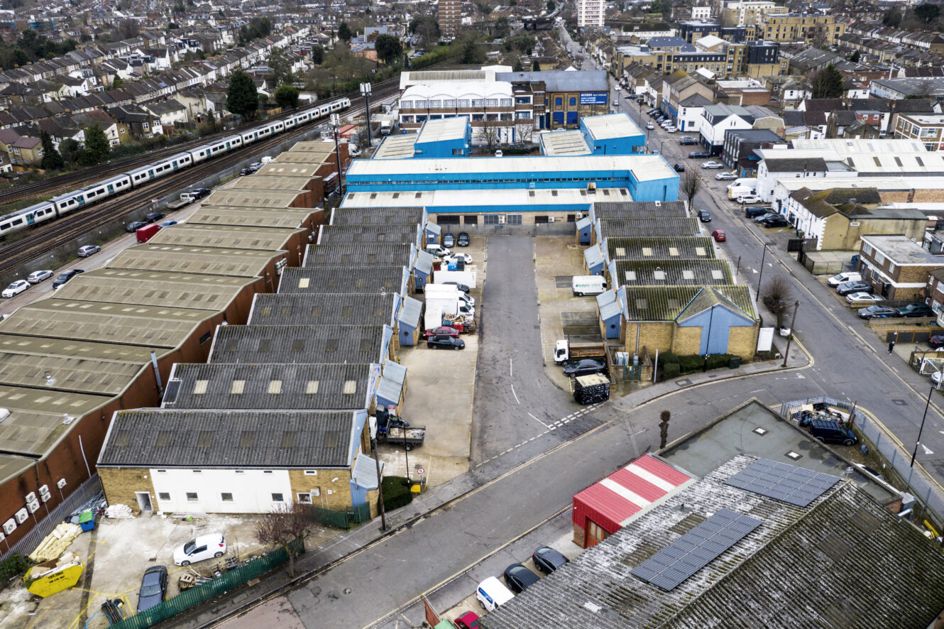 Aerial view of an industrial estate with multiple warehouse buildings, parked vehicles, and a train line running parallel to the site. Residential and commercial buildings surround the area.