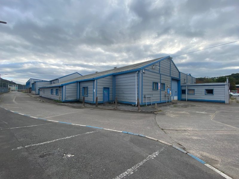 A large, light blue industrial warehouse building with metal siding, multiple entrances, and a mostly empty asphalt lot under a cloudy sky.