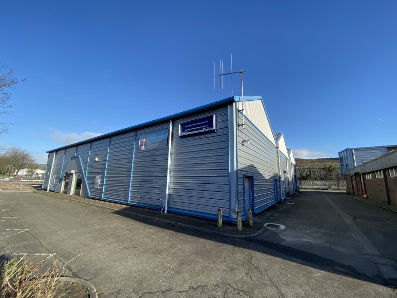 A large blue and gray metal industrial warehouse building with a sign above the door, surrounded by an empty parking lot under a clear sky.