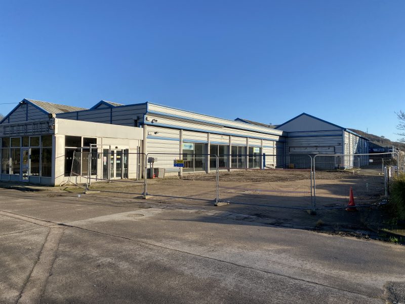 A vacant commercial building with large windows and metal siding is surrounded by metal fencing on a clear day.