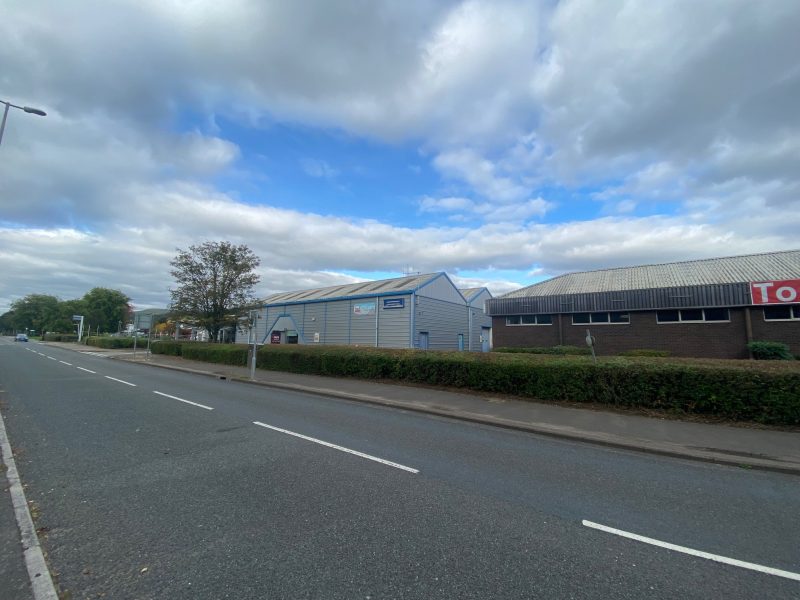 A quiet, empty street runs alongside industrial buildings with hedges and trees under a partly cloudy sky.