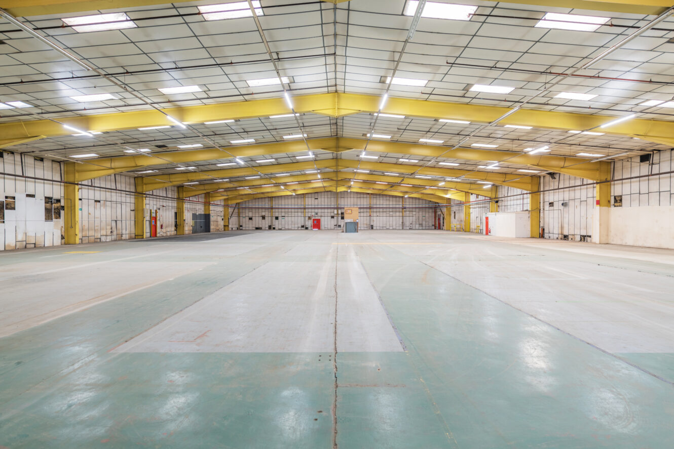Spacious, empty warehouse interior with high ceilings, yellow steel beams, overhead lighting, and a concrete floor.