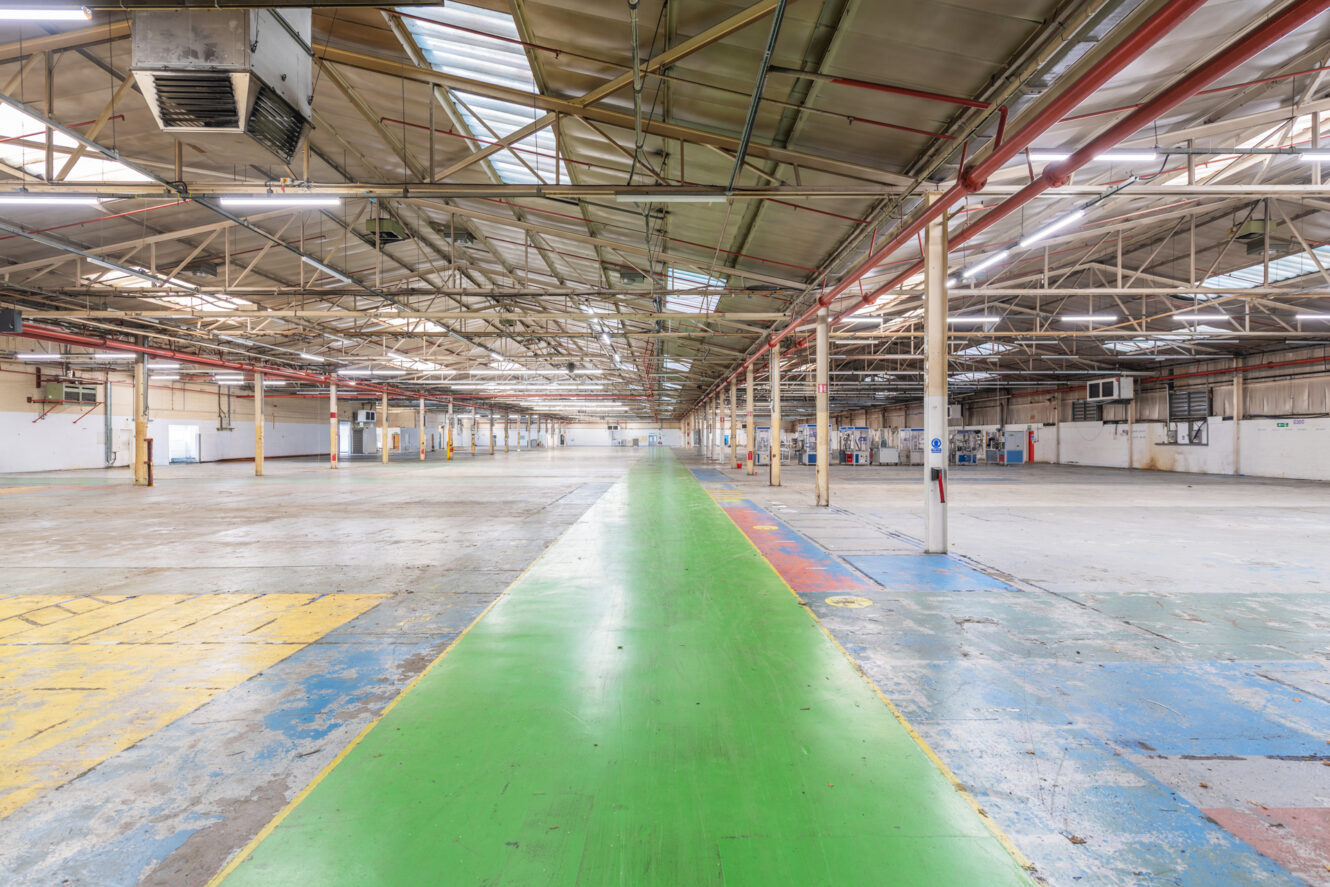 Large empty industrial warehouse with high ceilings, exposed beams, overhead lighting, and a green walkway running through the center of the worn concrete floor.