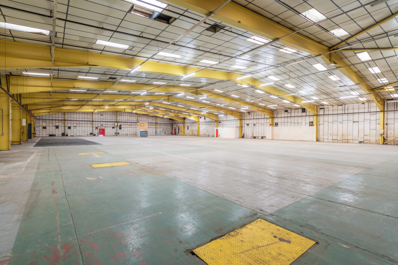Spacious, empty industrial warehouse with yellow steel beams, large overhead lights, and a worn concrete floor.