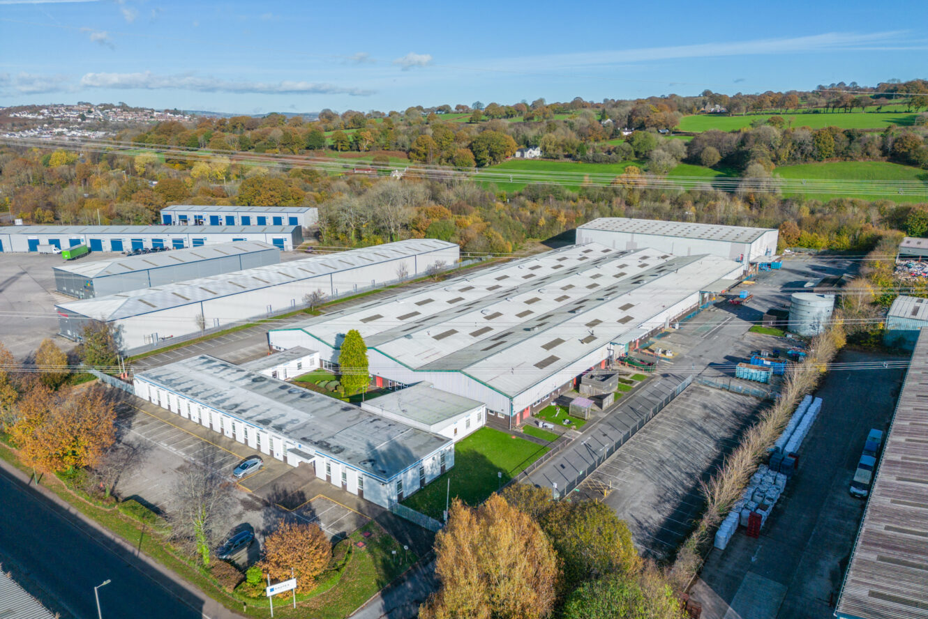 Aerial view of a large industrial warehouse complex surrounded by trees, roads, and adjacent industrial buildings, with grassy fields in the background.