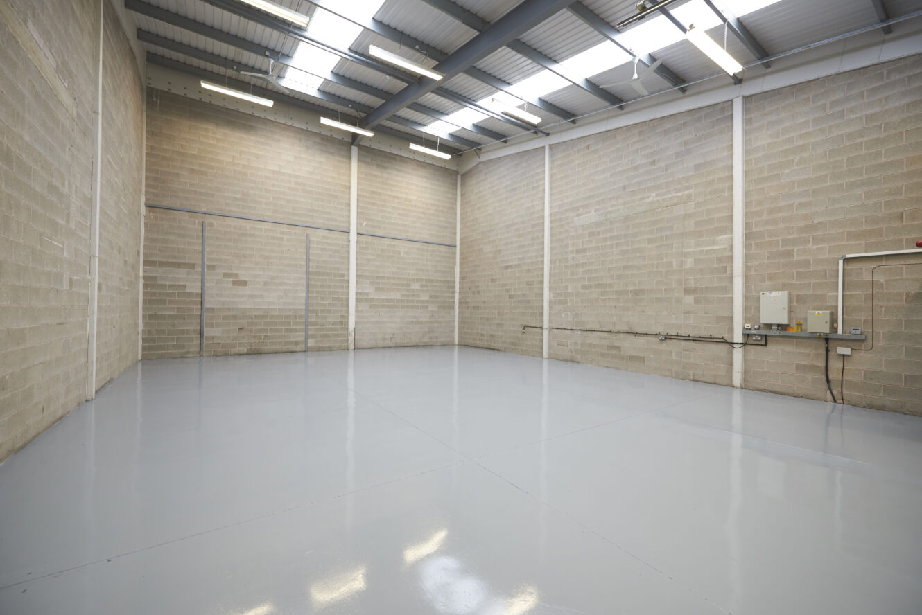 Empty industrial warehouse with concrete block walls, polished gray floor, high ceiling, skylights, and fluorescent lighting. Electrical panels are visible on the right wall.