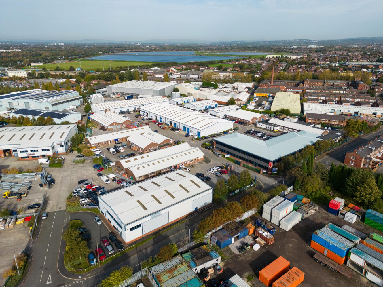 Aerial view of an industrial estate with multiple warehouses, parked vehicles, shipping containers, and surrounding greenery under a partly cloudy sky.