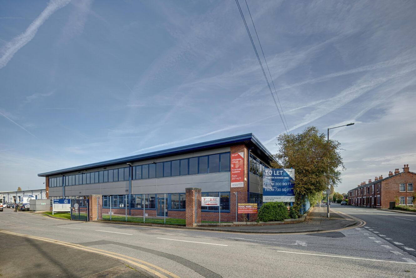 A modern two-story commercial building with To Let signs on the corner of a quiet street, surrounded by brick houses and clear skies overhead.