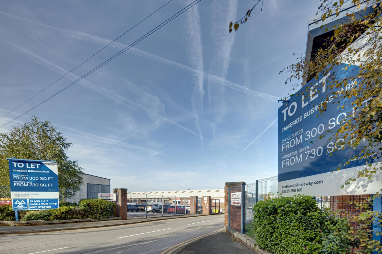 Entrance to Tameside Business Park with signs advertising industrial units to let, blue sky overhead, and trees on the side.