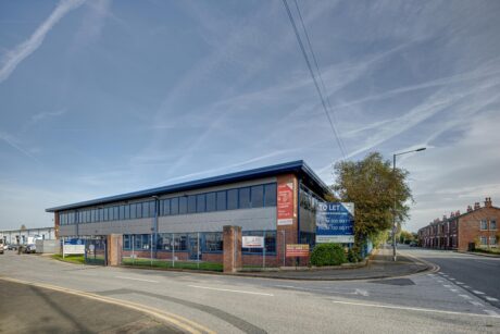 A modern commercial building with large windows on a street corner, featuring To Let signs and surrounded by residential houses and clear skies.