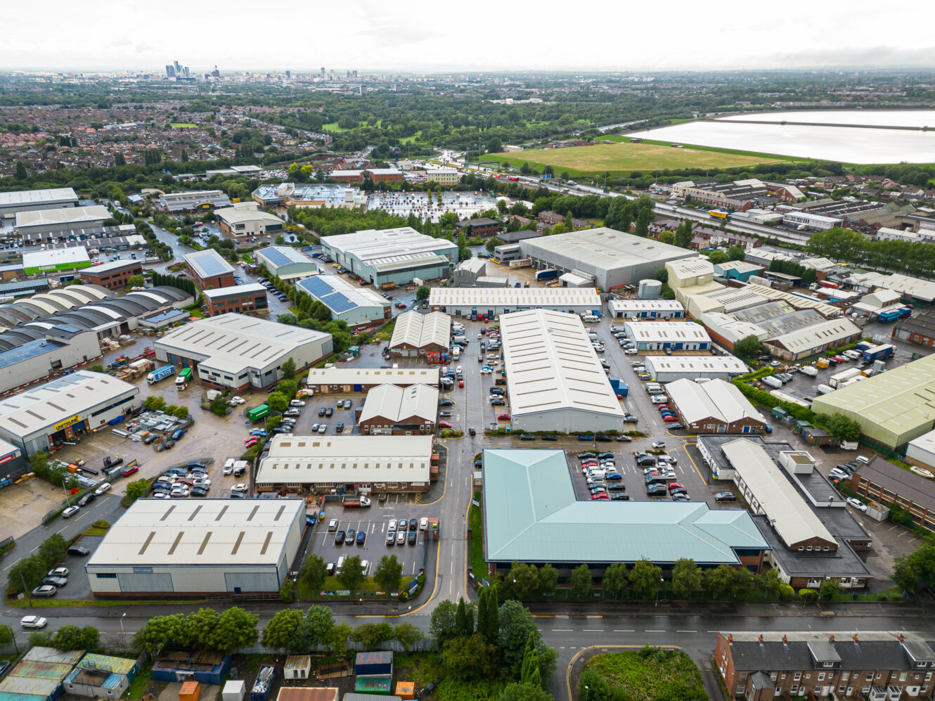 Aerial view of an industrial estate with multiple warehouses, parking lots filled with vehicles, and green fields in the background under a cloudy sky.