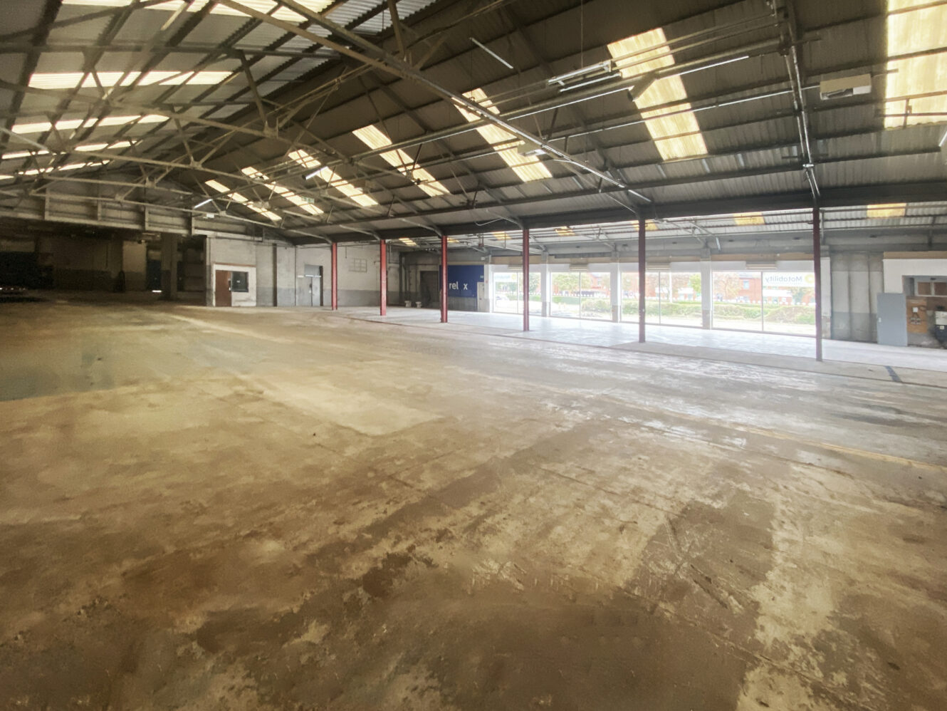 Large, empty warehouse interior with concrete floor, exposed steel beams, skylights, and open shutter doors letting in natural light.