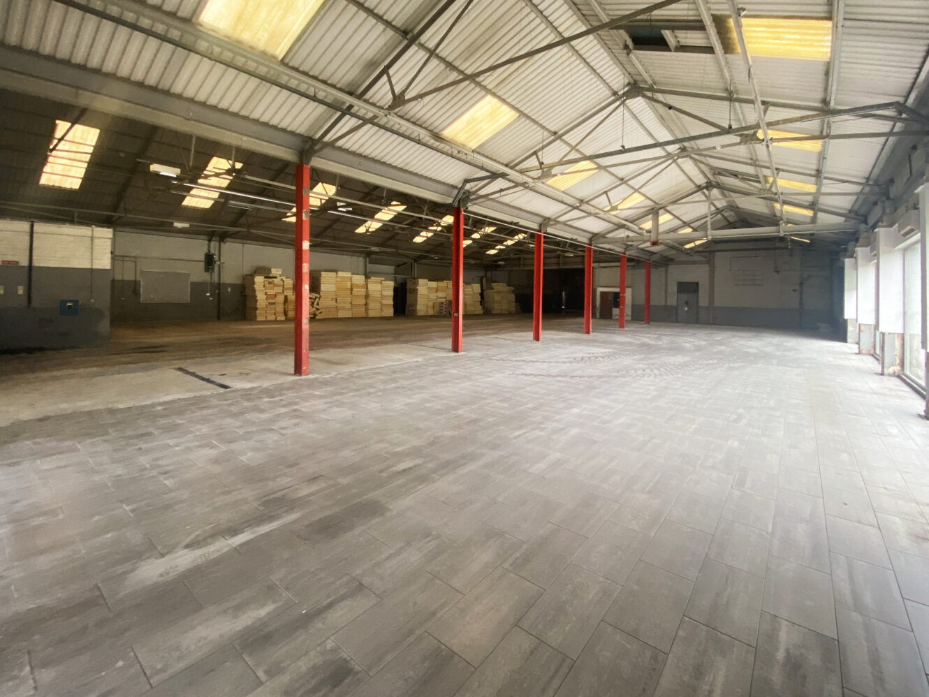 Large empty warehouse with tiled flooring, high ceiling, red support beams, stacks of pallets, and natural light from windows on the right.