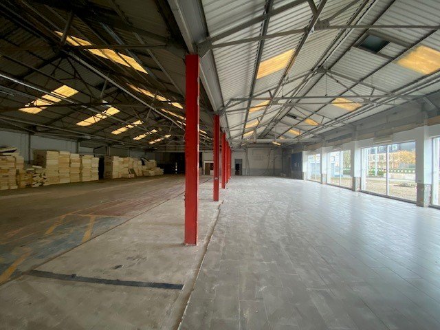 Large empty warehouse interior with red columns, stacked wooden pallets on the left, and tiled flooring on the right near large windows.