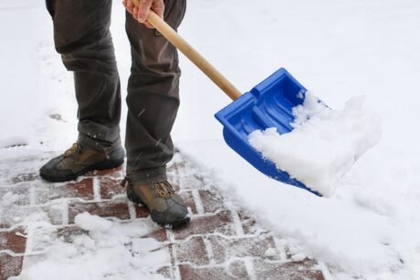 Person shoveling snow from a brick walkway with a blue plastic snow shovel. Only the lower half of the person is visible.
