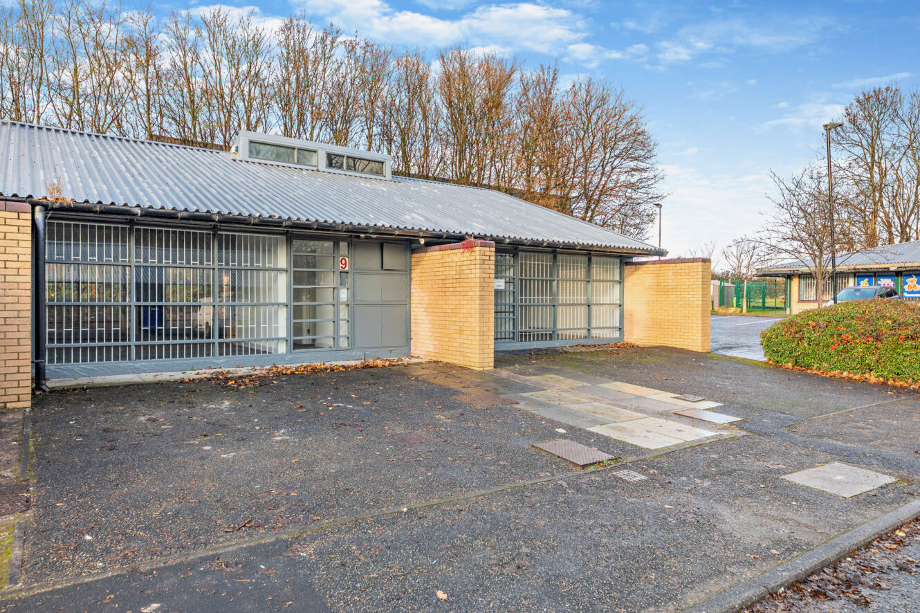 Single-story building with barred windows and brick walls, surrounded by trees and pavement under a partly cloudy sky.