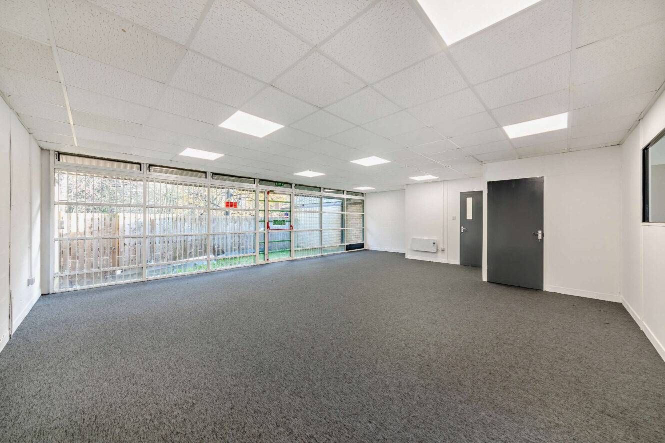 Empty room with gray carpet, white walls, a dark door, fluorescent ceiling lights, and large windows along one side showing a fenced outdoor area.