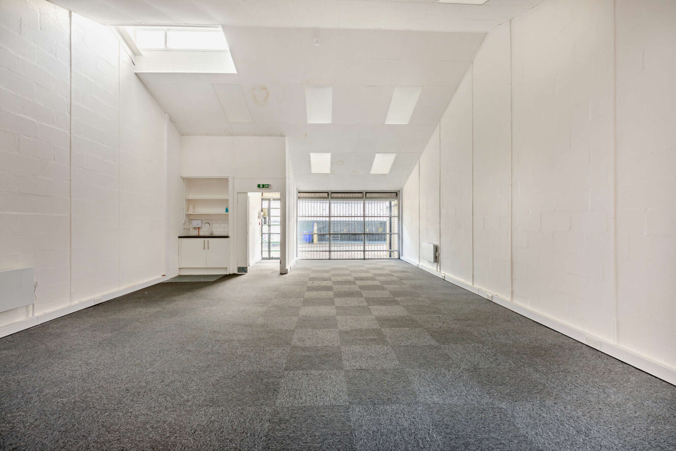 Spacious, empty commercial or office space with high ceiling, white walls, grey carpet tiles, skylights, and a small kitchenette in the corner.