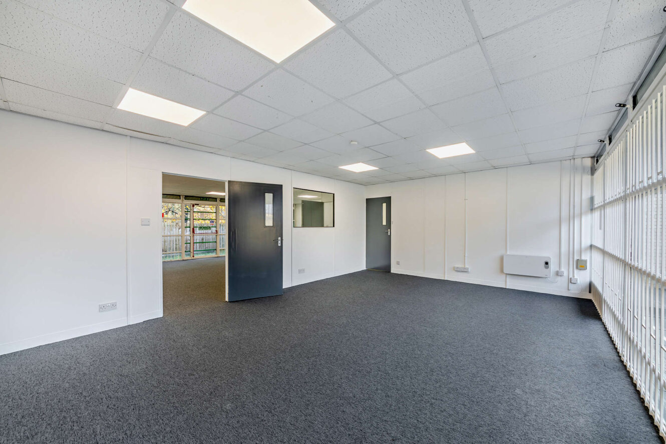 Empty office room with gray carpet, white walls, ceiling lights, large window with vertical blinds, and two gray doors leading to adjacent rooms.