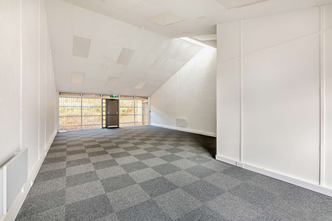 Empty room with white walls, checkered gray carpet, slanted ceiling, large windows, and a glass door leading outside.
