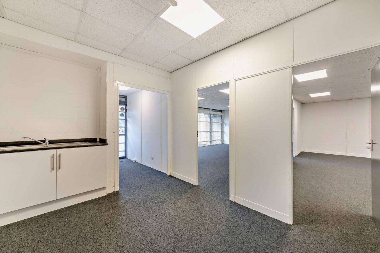 Interior of an empty office space with white walls, gray carpet, ceiling lights, a kitchenette area, and multiple open doorways leading to other rooms.