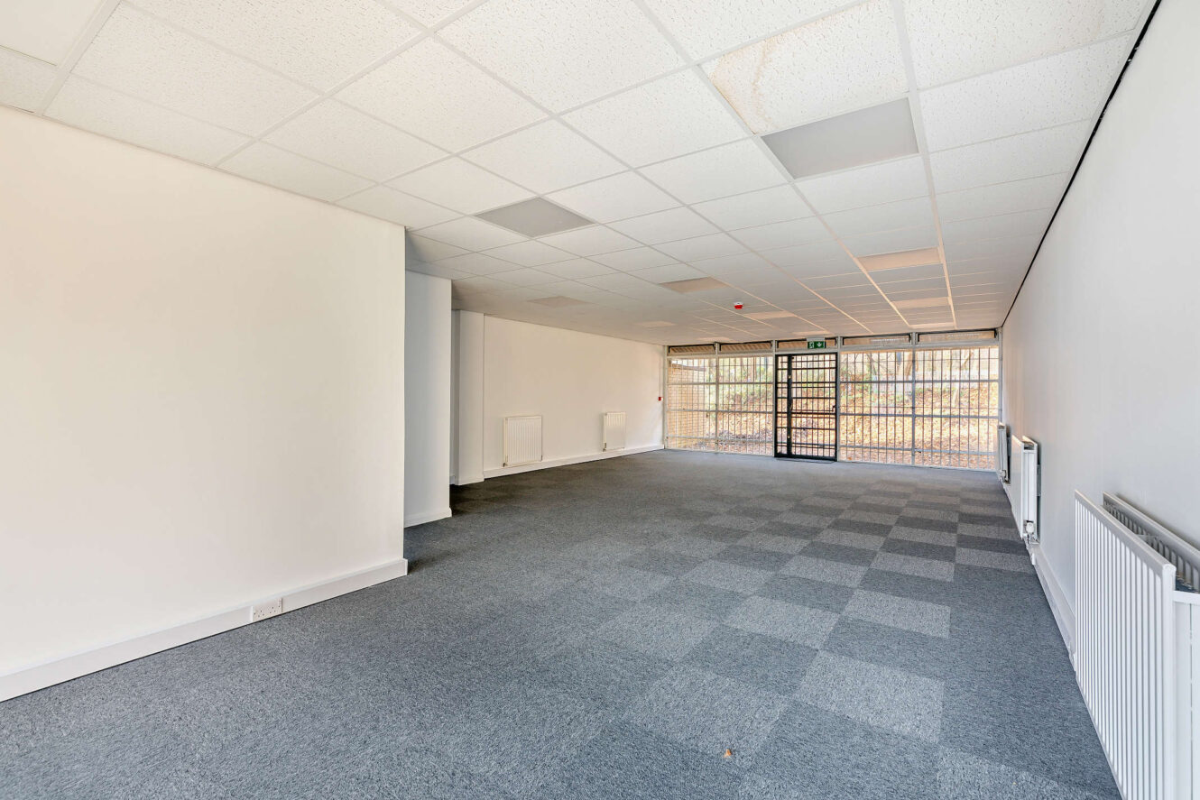Empty room with gray carpet tile flooring, white walls, and a grid-patterned drop ceiling. Large windows with security bars and a glass door are at the far end.