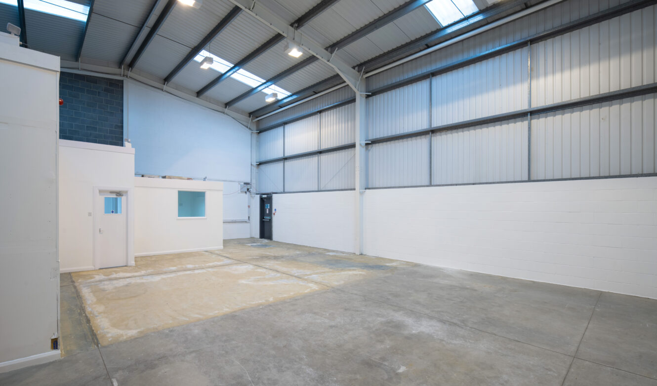 Empty industrial warehouse with high ceilings, concrete floor, white painted walls, large windows, and a small enclosed office area.