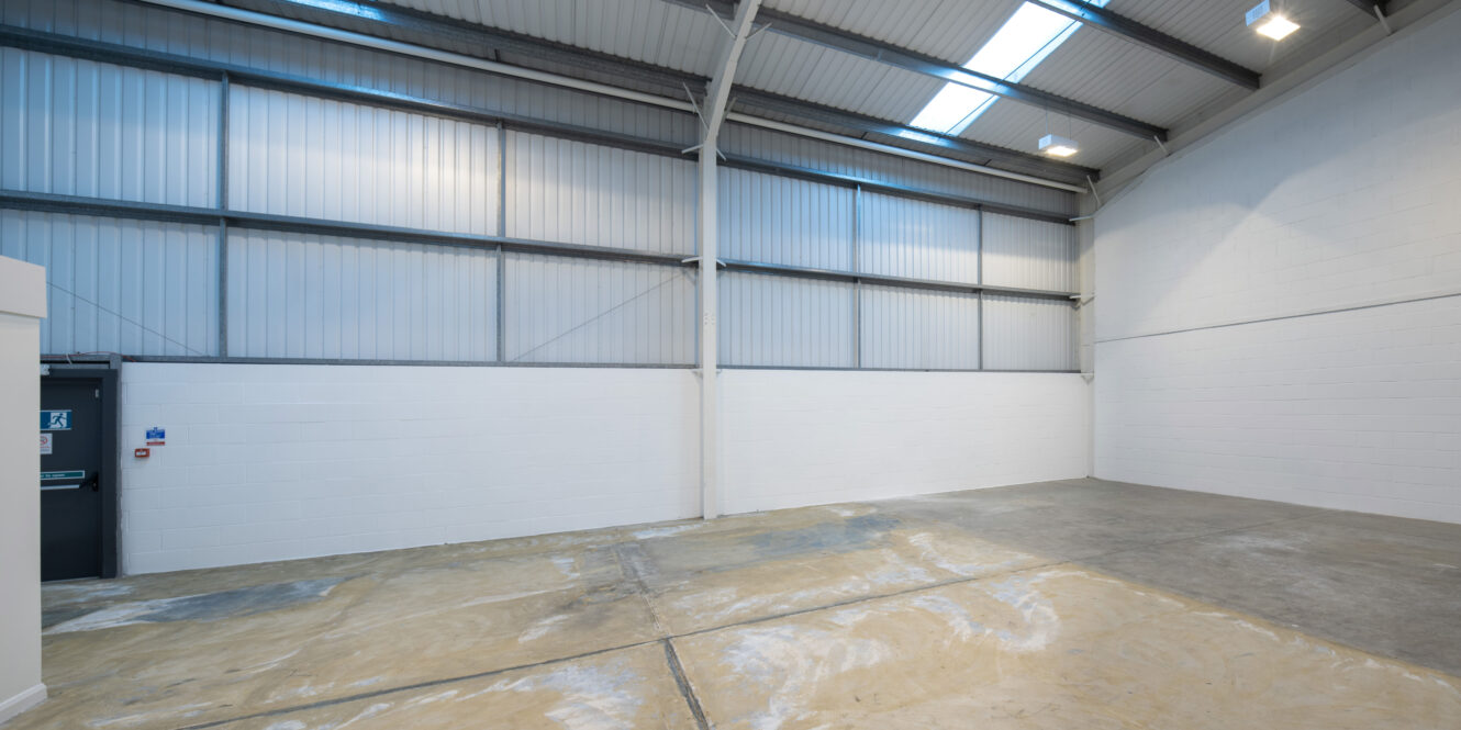 Empty industrial warehouse interior with concrete floor, high ceiling, metal beams, and white painted brick walls. Natural light enters through skylights and windows on the upper walls.