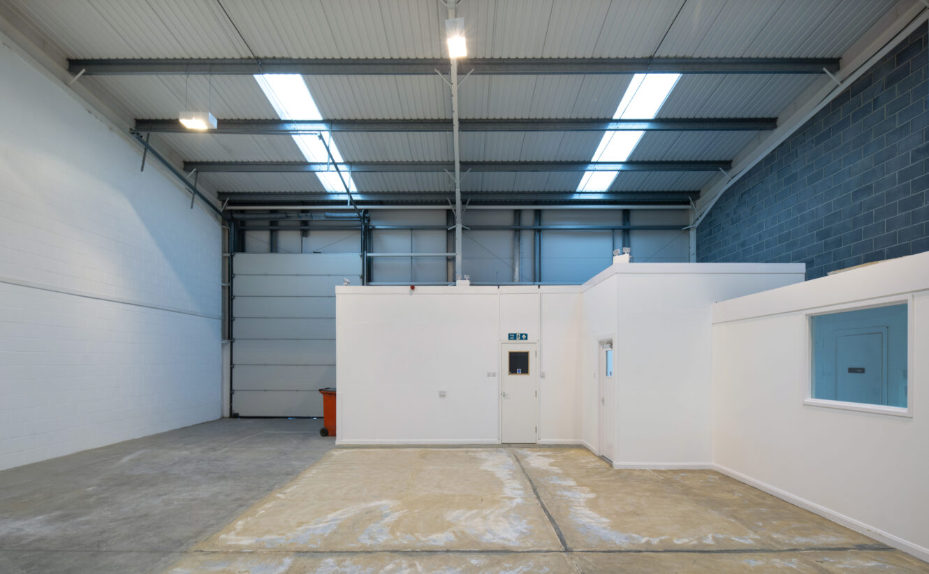 Interior of an empty industrial warehouse with high ceilings, large overhead lights, and a small white office structure inside.