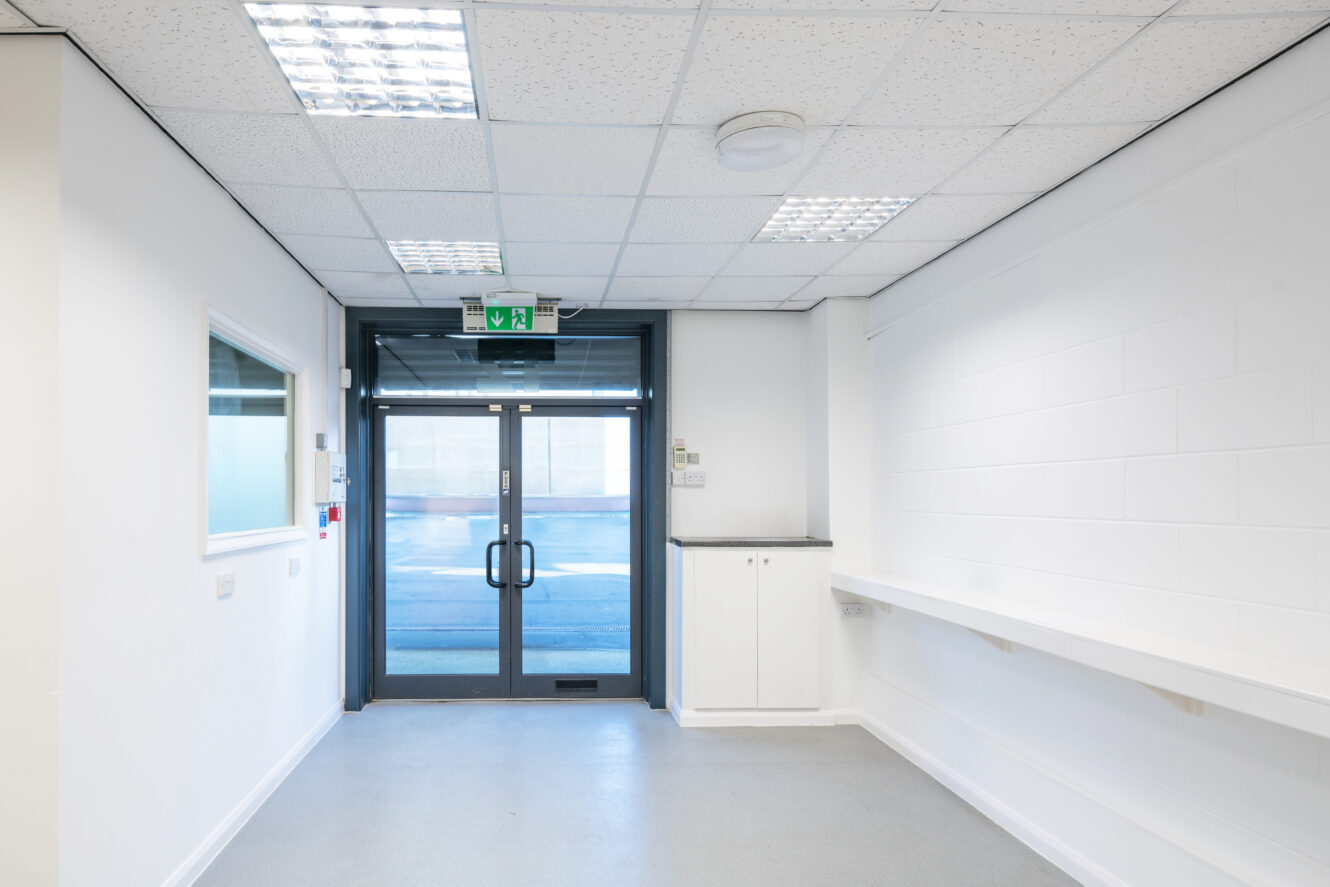 An empty, well-lit hallway with white walls, a long counter, overhead lights, and double glass exit doors at the end.