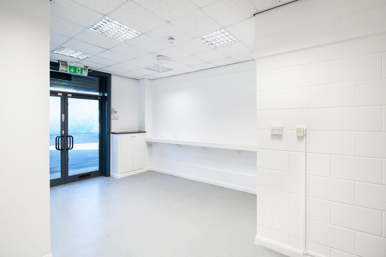 Empty, well-lit room with white walls, gray flooring, a glass double door, built-in shelf, storage cabinet, and ceiling fluorescent lights.