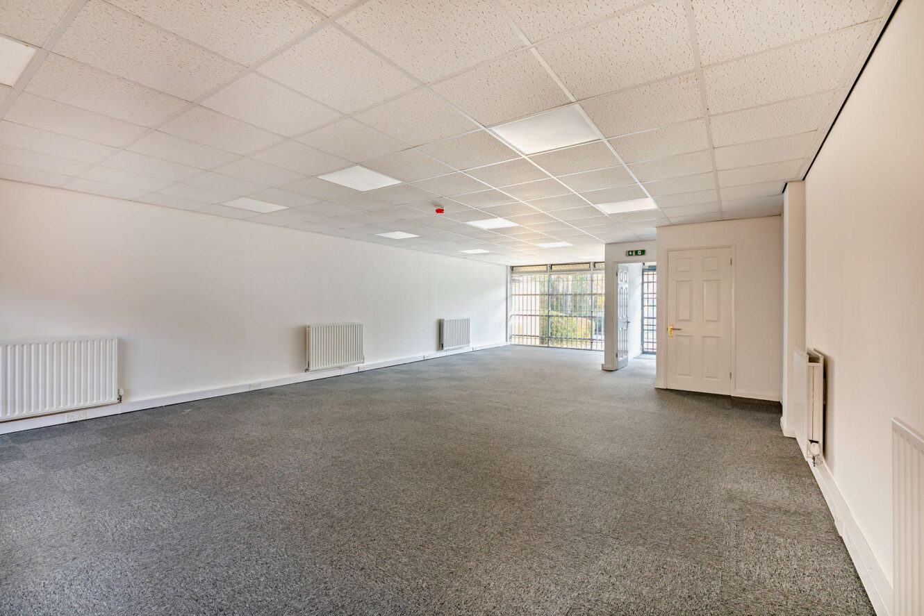 Empty office space with gray carpet, white walls, ceiling tiles, radiators, and large windows with security bars near the entrance door.