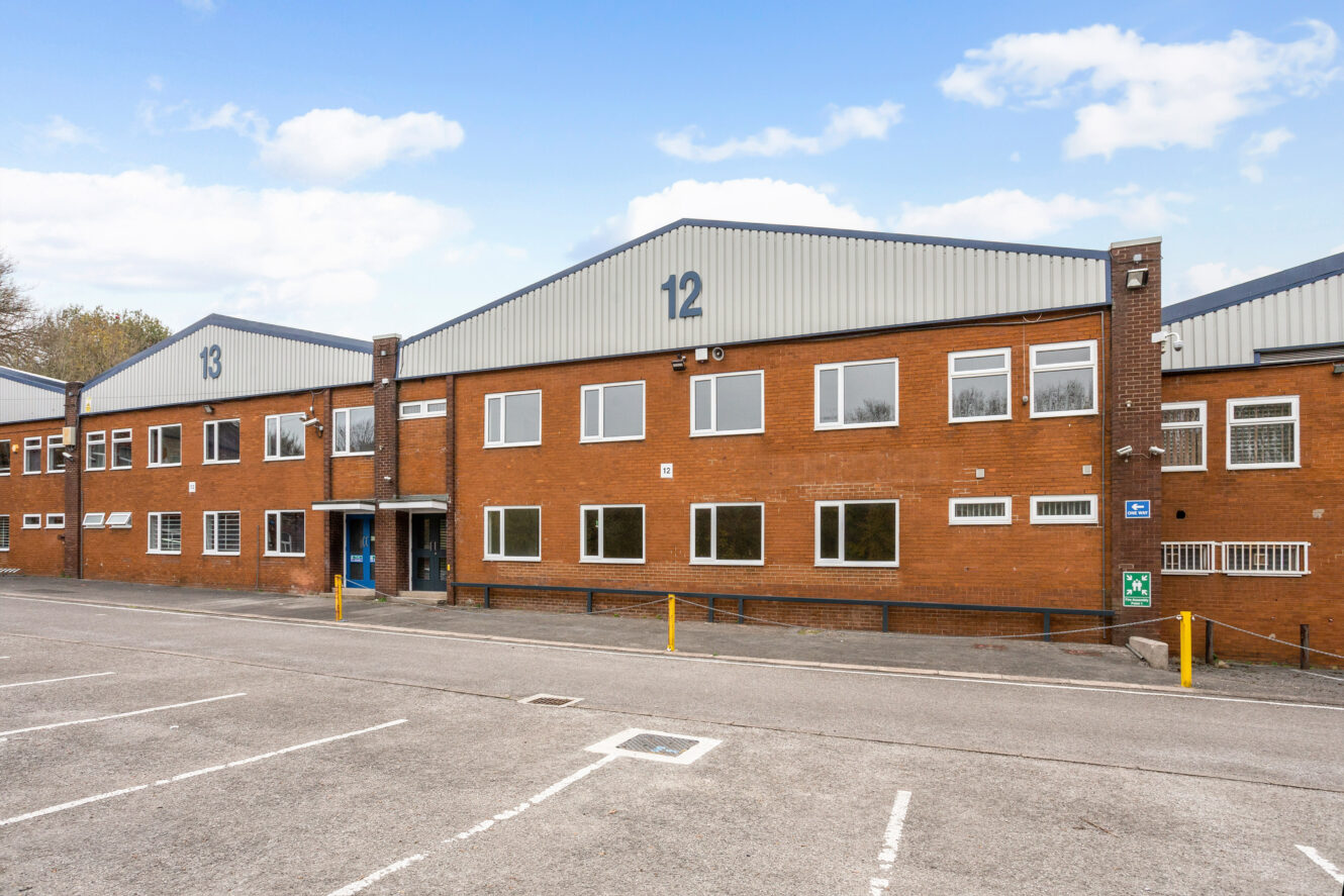 A two-story industrial building labeled 12 with a brick facade, large windows, and an empty parking lot in front under a partly cloudy sky.