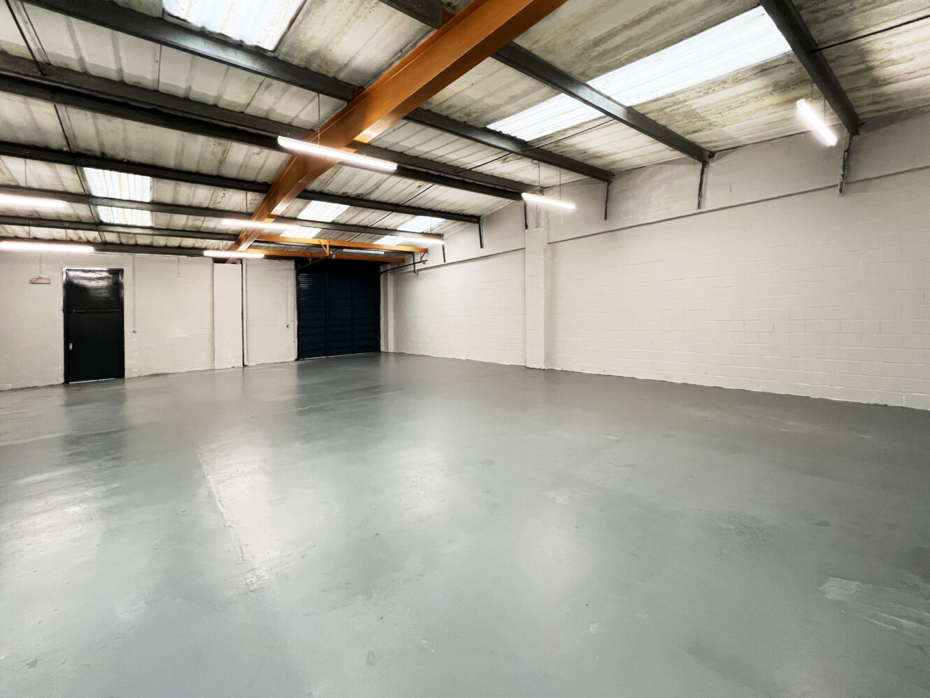 Empty industrial warehouse with smooth gray floor, white painted brick walls, exposed beams, and overhead fluorescent lighting.