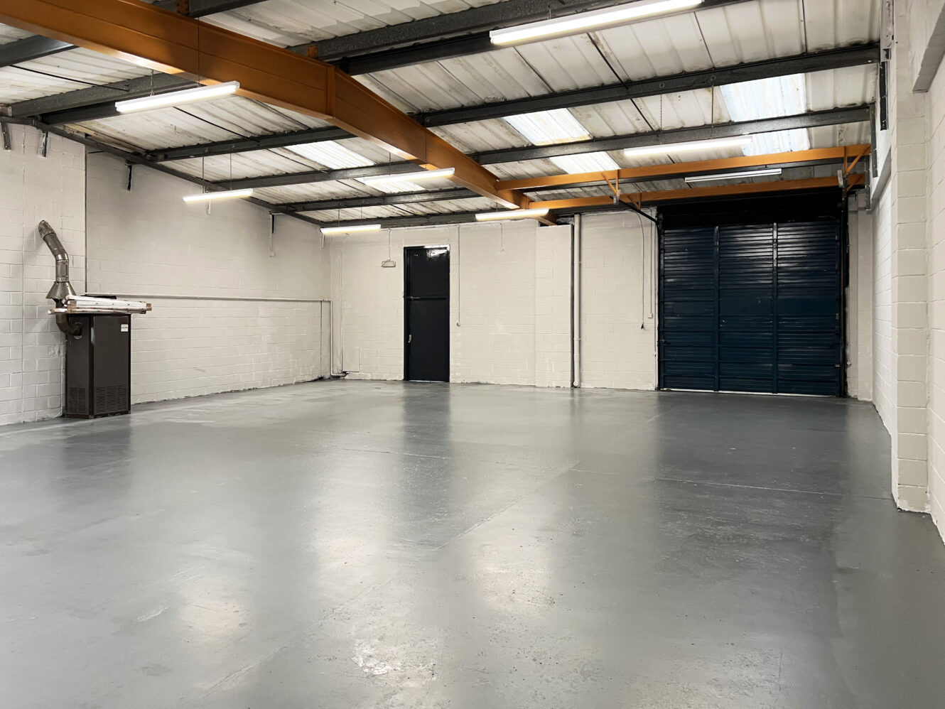 Empty industrial warehouse with concrete floor, white brick walls, exposed beams, fluorescent lights, a black door, and a large closed roller shutter.