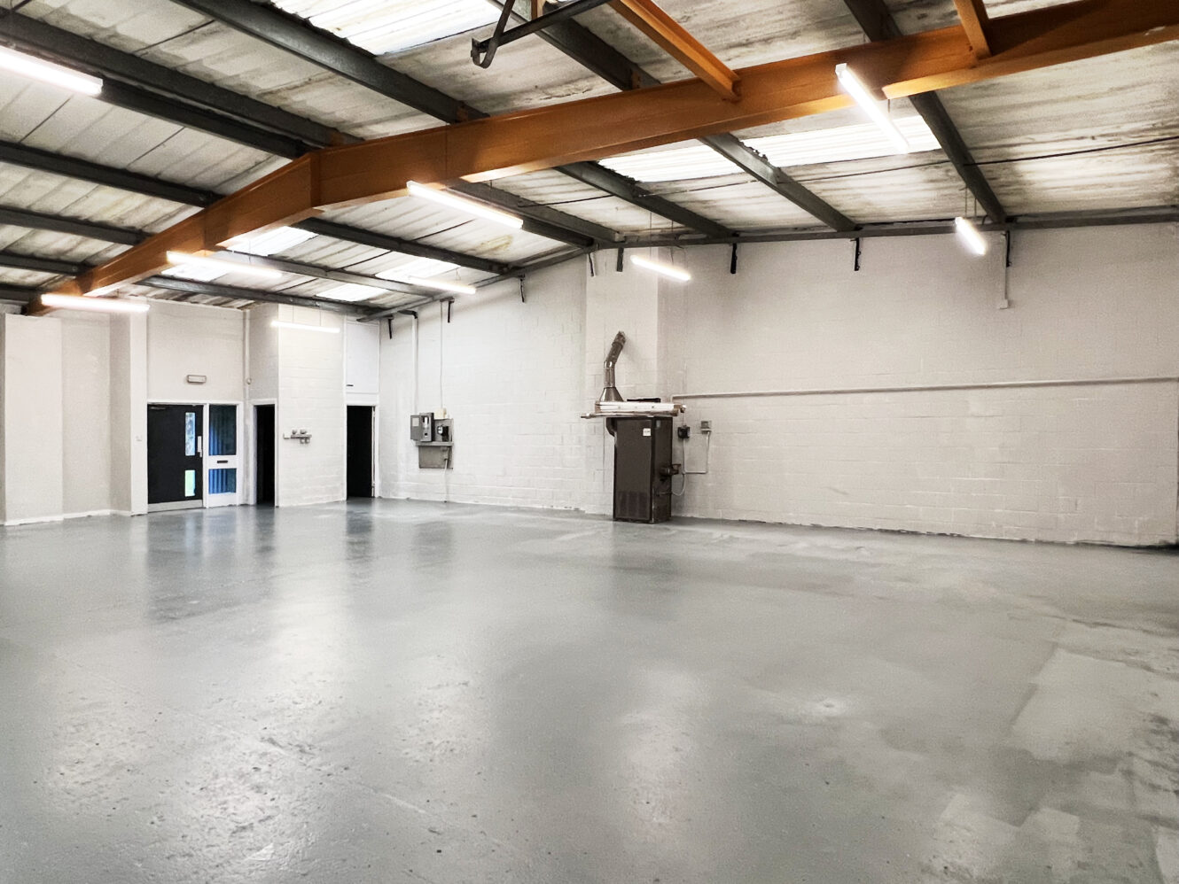 Empty industrial warehouse with a concrete floor, exposed beams, white brick walls, fluorescent ceiling lights, and a few utility panels on the far wall.