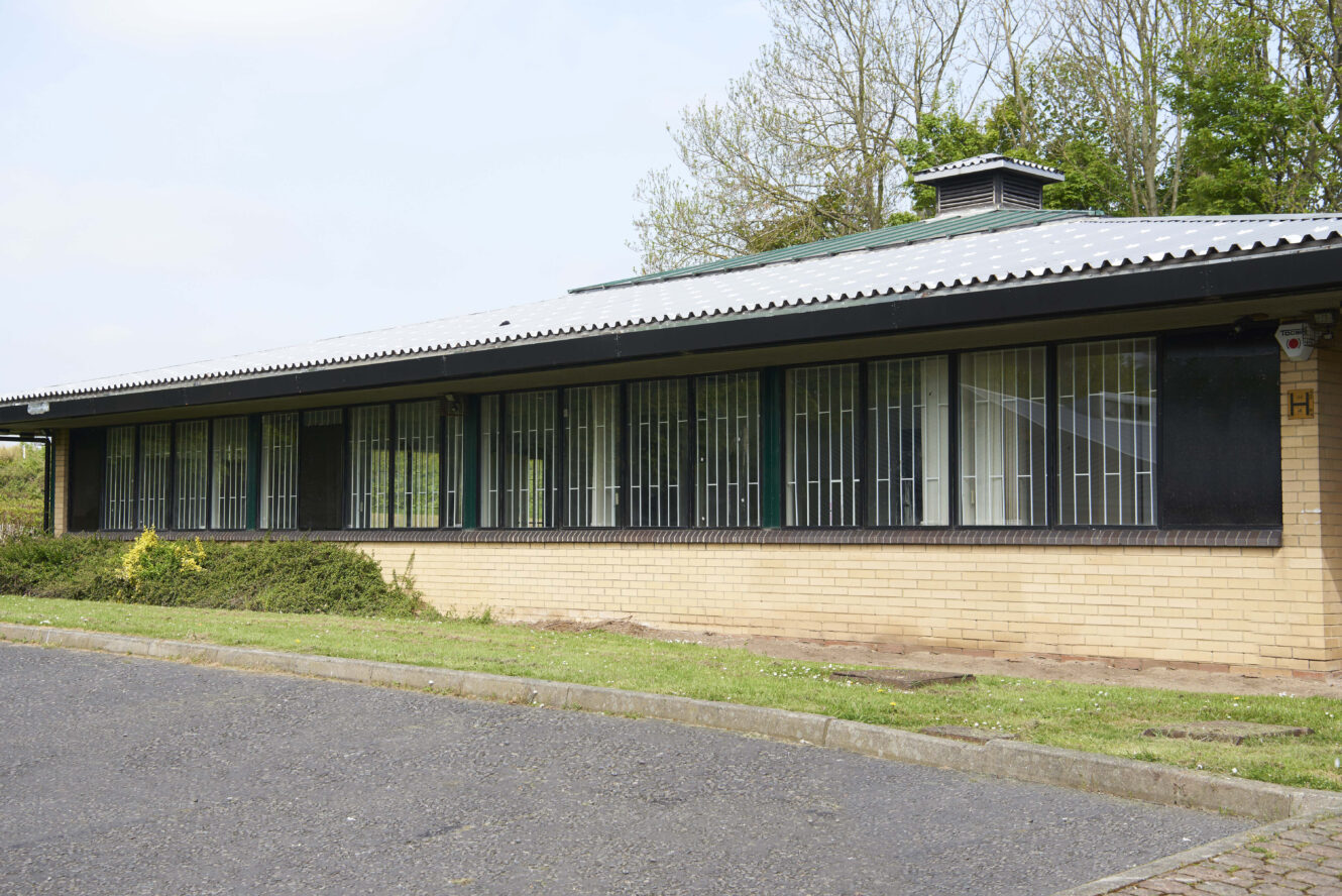 Single-story brick building with long vertical windows, surrounded by grass, shrubs, and trees, under a light blue sky.