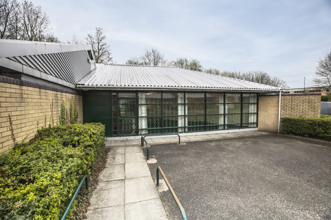 Single-story brick building with large windows and a corrugated metal roof, surrounded by shrubs, a paved path, and an empty parking area in front.