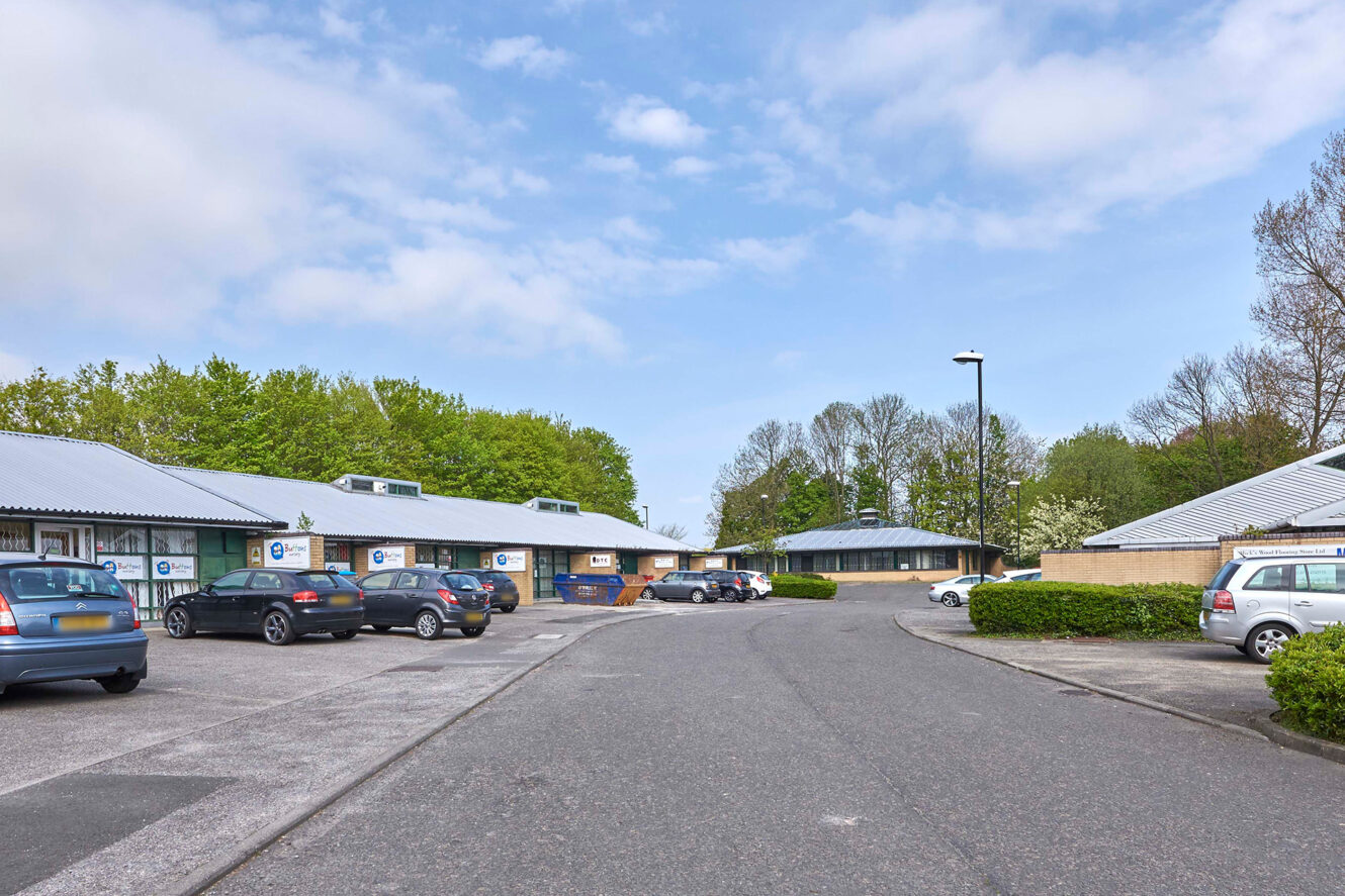 A parking lot with several cars in front of low, single-story commercial buildings with signs; trees and a partly cloudy sky in the background.